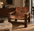 A rustic mid-century modern accent chair with a dark brown wooden frame and a distressed brown leather sling seat and backrest featuring a black printed cowboy on horse design, set in a warm, sunlit living room.