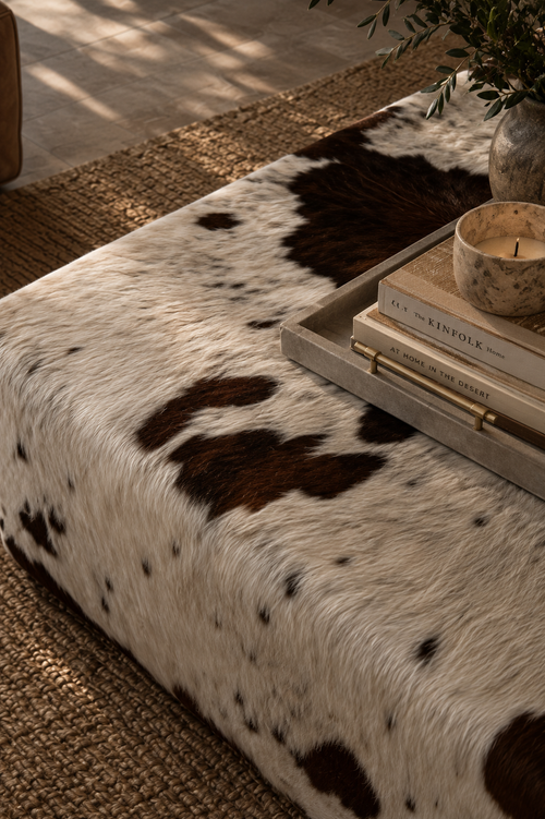 A cowhide ottoman or bench with a black-and-white spotted pattern, styled in a warm interior setting with a tray, books, candle, and plant on top.