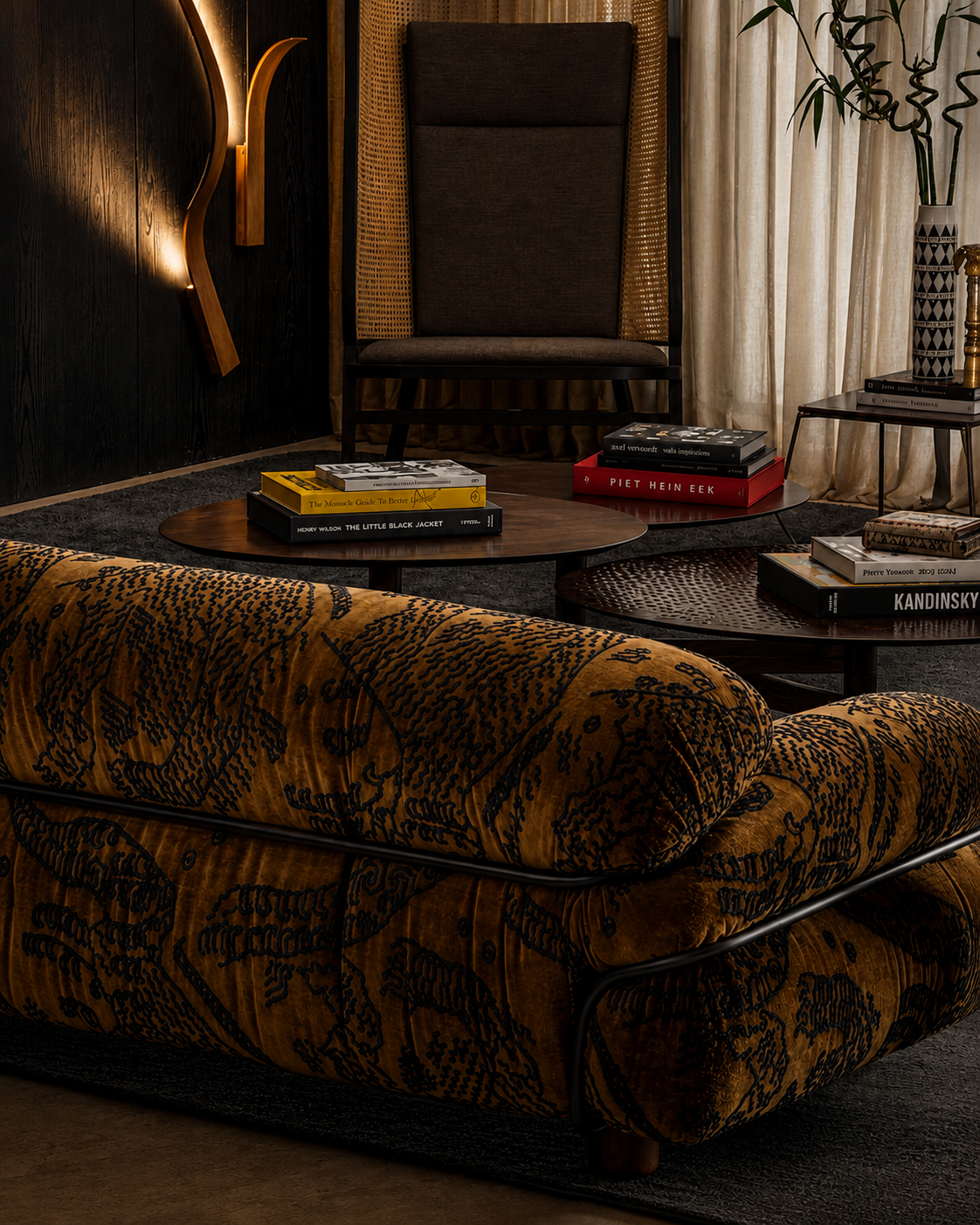 A close-up of a luxurious velvet sofa in mustard yellow with a black botanical pattern, set in a stylishly decorated living room with a dark wood chair, coffee tables, and books.