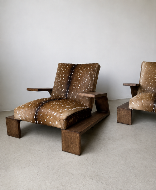 A mid-century modern wooden lounge chair with brown and white spotted animal-print upholstery, shown in a minimalist room with a second matching chair partially visible.